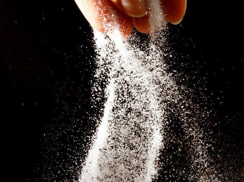 Manos tiran sal sobre un fondo negro. Aprende sobre los síntomas del exceso de sal en el cuerpo.