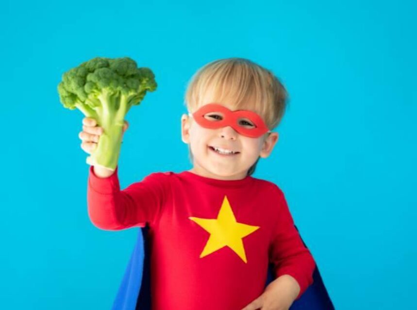 Un niño vestido de súper héroe sostiene un brócoli en su mano derecha.