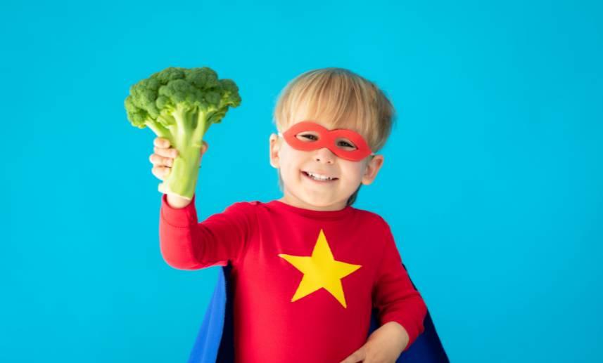 Un niño vestido de súper héroe sostiene un brócoli en su mano derecha.