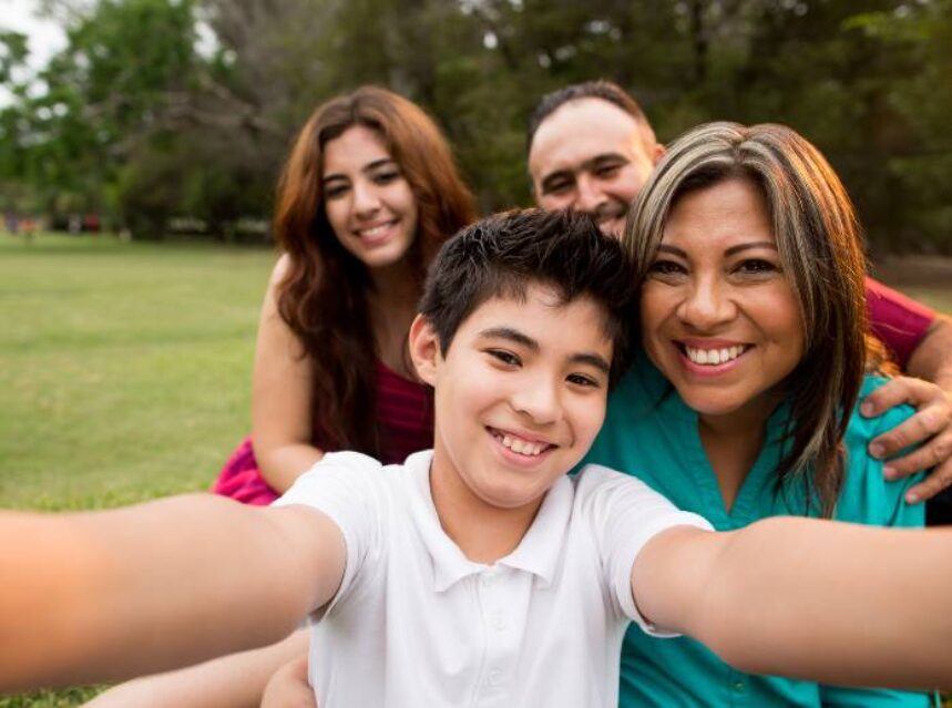Una familia de 4 integrantes sonríen a la cámara en un verde pasto de un parque.