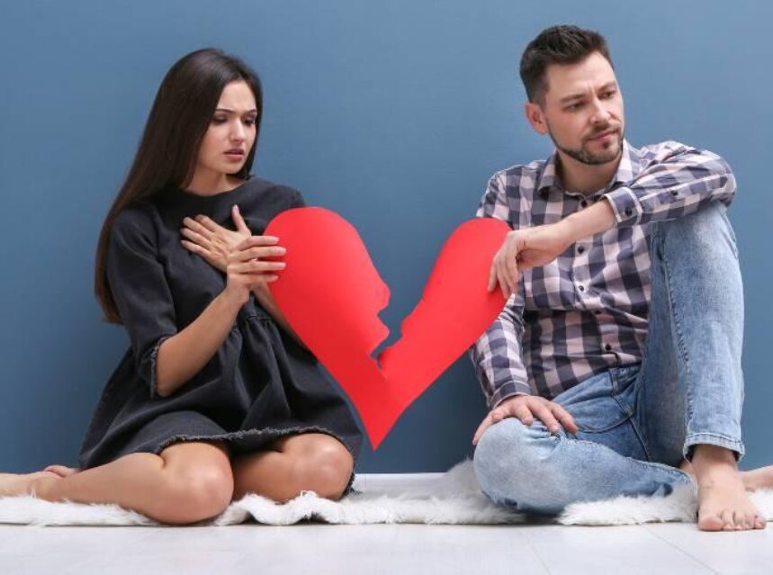 Una mujer y un hombre sentados toman cada uno con una mano un corazón rojo de papel que se está rompiendo. Ambos tienen un semblante de dolor en sus rostros.