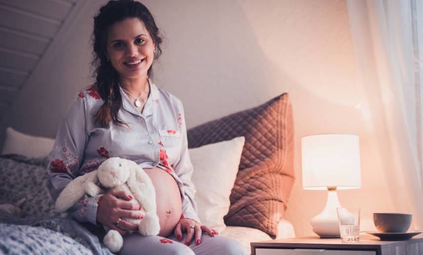 Mujer embarazada en pijama sostiene un conejo de peluche, está sentada sobre una cama.