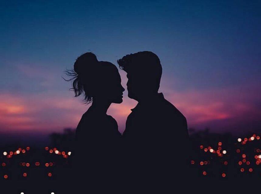 Contorno de una pareja frente a frente en sombra, con un paisaje de puesta de sol casi anocheciendo al fondo y luces de una ciudad.