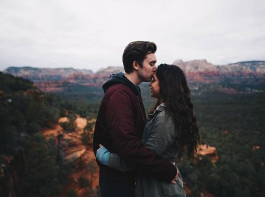 Hombre y mujer abrazados, con un fondo de montañas. El hombre besa cariñosamente la frente de la mujer.