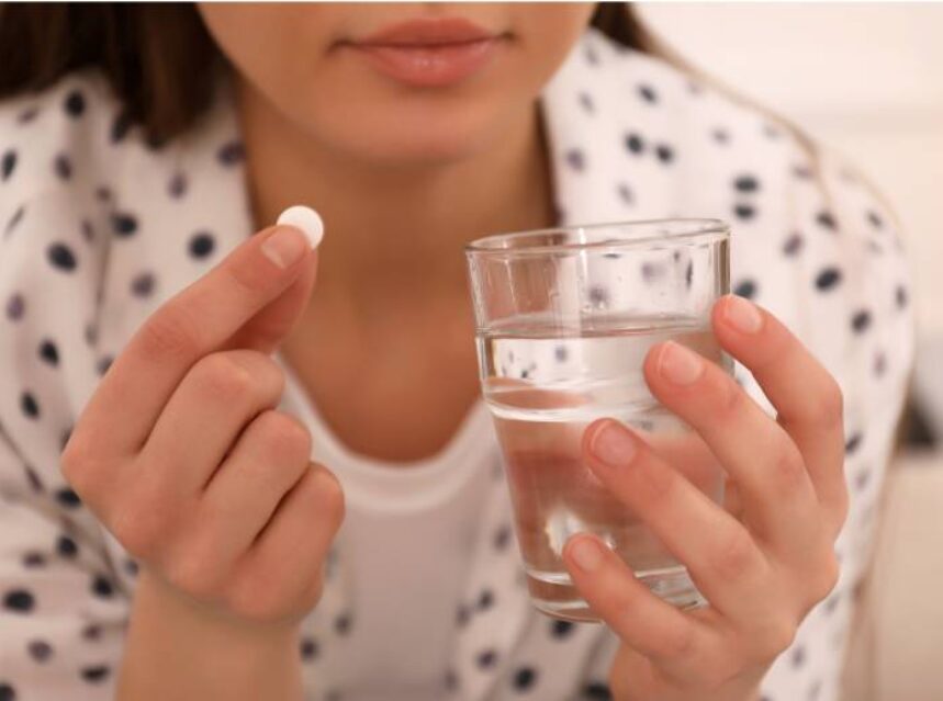 Una mujer sostiene en la mano derecho una pastilla y en la mano izquierda un vaso con agua.