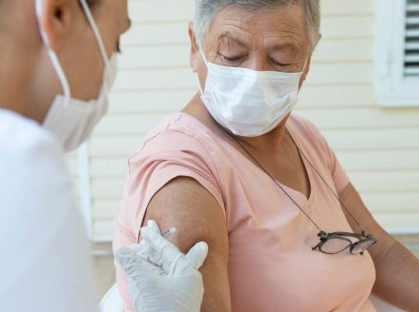 Mujer con cubrebocas recibe vacuna en brazo derecho a través de otra mujer de los servicios de salud.