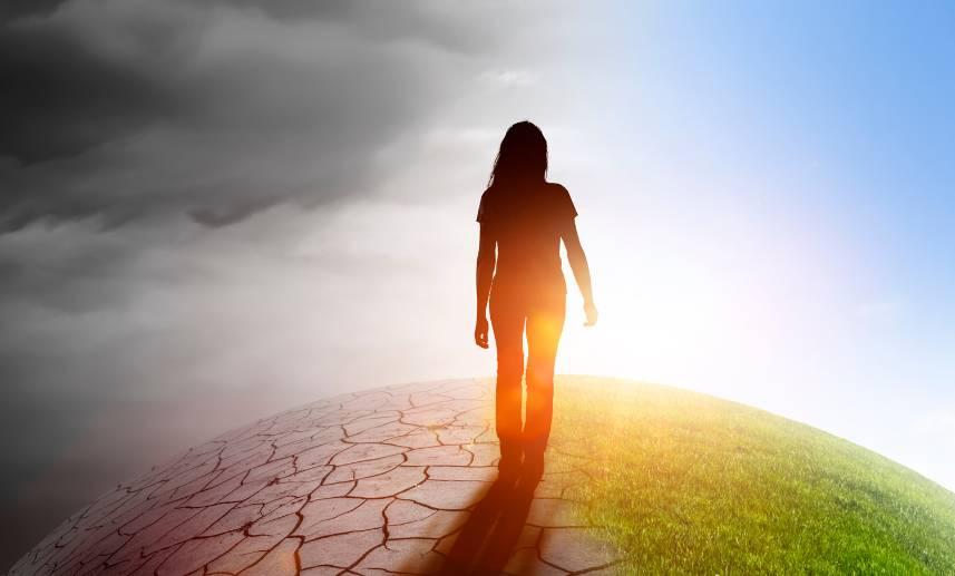 Mujer camina por un paisaje que en una mitad es tierra erosionada y un cielo nublado, por la otra se ve una superficie verde con un cielo azul.