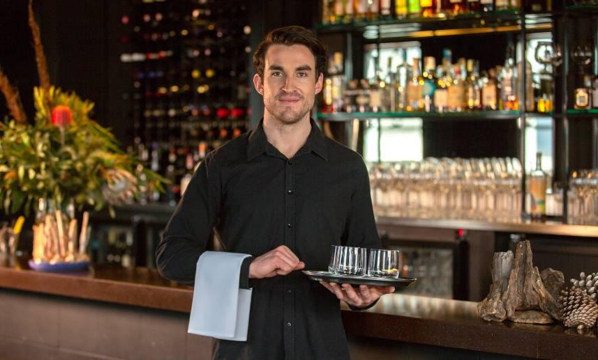 Joven hombre sostiene una charola con tragos de bebidas, en lo que parece un bar nocturno.