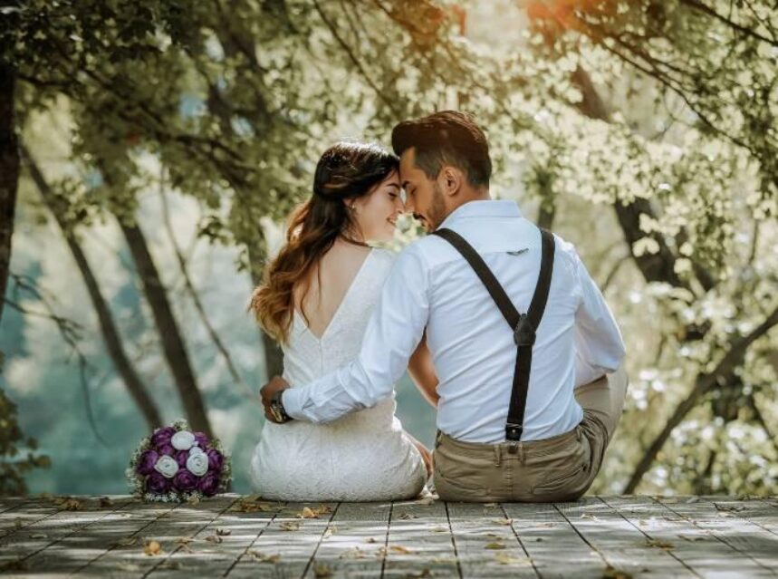 Una pareja en su boda permanece sentada en un muelle, mientras se abrazan y hacen cariños. Al fondo hay flores.