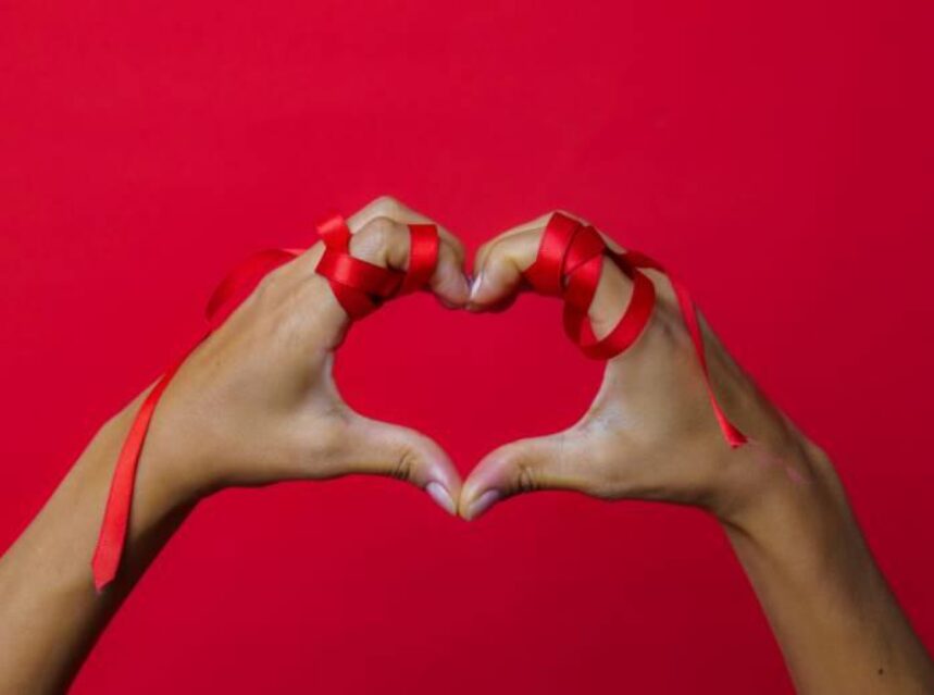 Un par de manos firman un corazón, les sobresale un listón color rojo, en un fondo también rojo en señal de lucha contra el VIH.