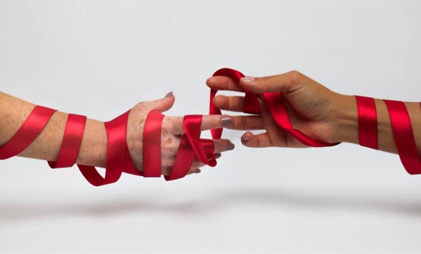 Dos manos a punto de tocarse sostienen, entrelazadas, un listón rojo, en señal de la sensibilización por el VIH.