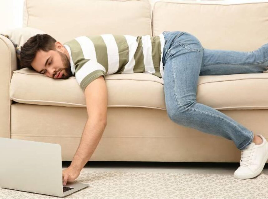 Hombre tirado boca abajo en su sofá, teclea sobre su laptop que está en el piso.