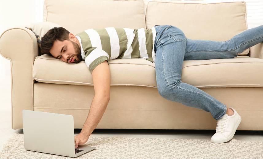 Hombre tirado boca abajo en su sofá, teclea sobre su laptop que está en el piso.