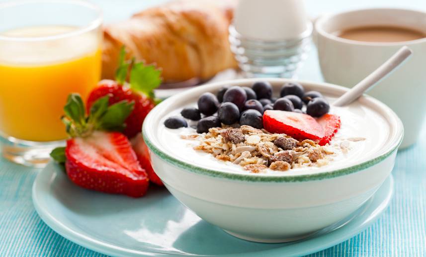 Un plato de yogourt con frutas y granola, al fondo, un cuernito y café con leche. Un desayuno completo.