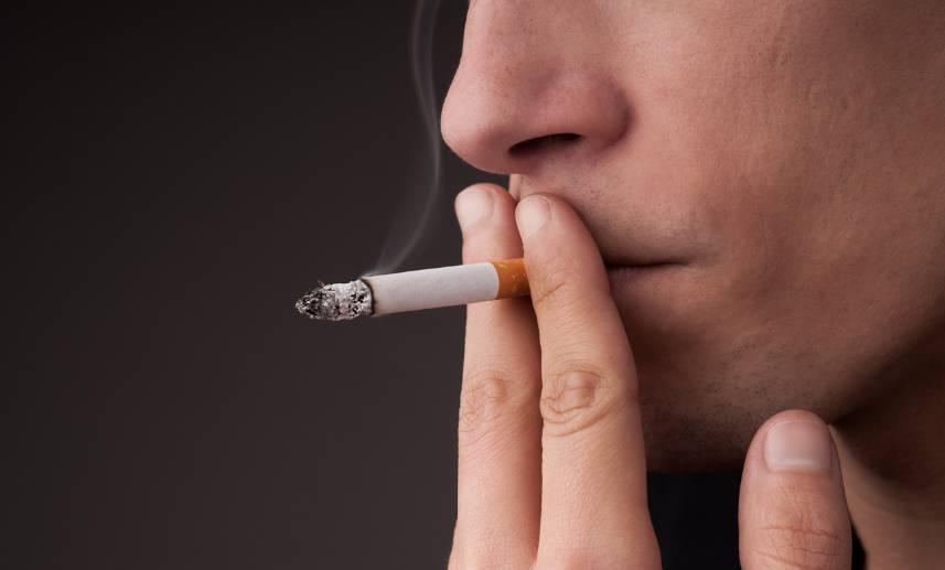 Rostro de persona que sostiene entre sus labios un cigarrillo, rodeado por sus dedos.