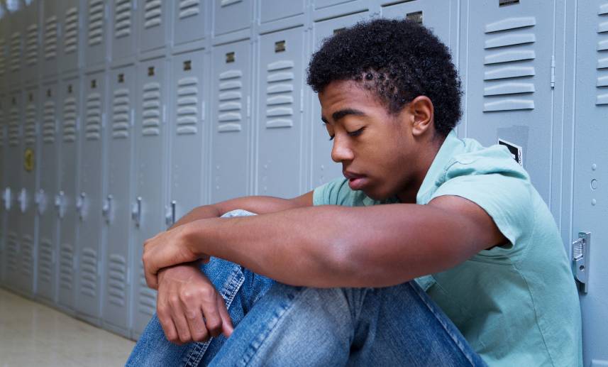 Joven sentado con la mirada hacía el suelo, justo en la zona de lockers de su centro educativo.