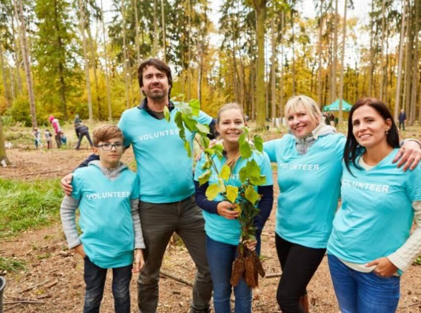 Grupo de 5 voluntarios en un bosque se abrazan mientras sonríen a la cámara y una de las personas sostiene un árbol para ser plantado.