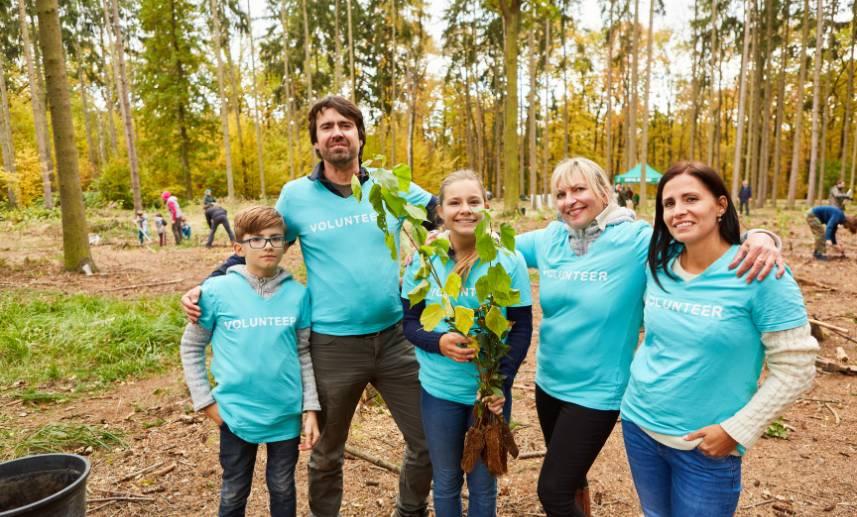 Grupo de 5 voluntarios en un bosque se abrazan mientras sonríen a la cámara y una de las personas sostiene un árbol para ser plantado.