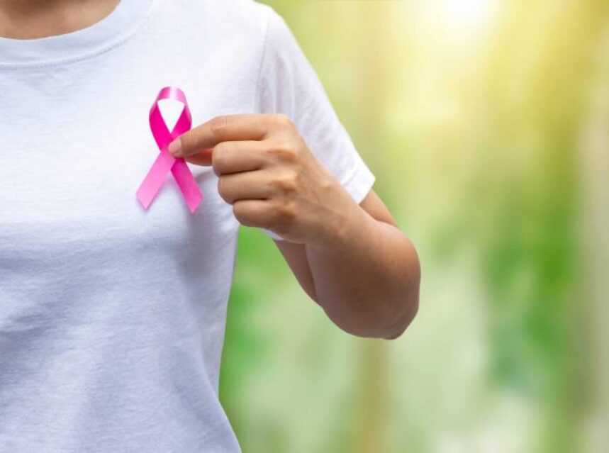 Parte superior del cuerpo de una mujer con playera blanca, sostiene con su mano izquierda a la altura del corazón un listón color rosa, símbolo de la prevención del cáncer de mama.