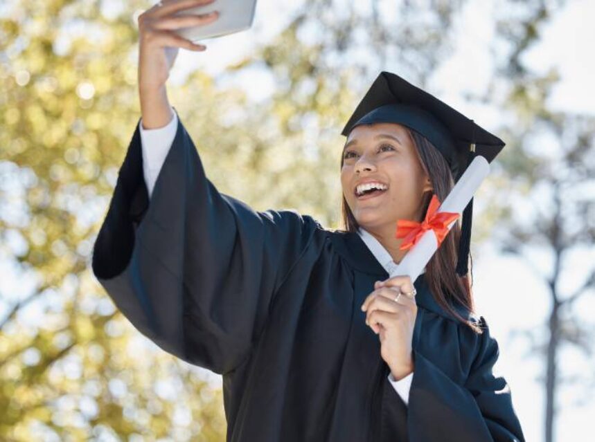 Una joven alegremente posa ante su celular para una selfie, porta en su mano izquierda su diploma, porta su toga y birrete.