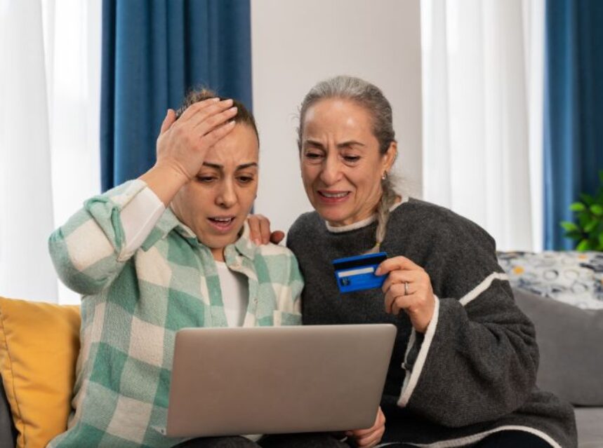 Dos mujeres frente a una pantalla de laptop miran con impresión mientras una de ella sostiene un plástico de tarjeta bancaria.