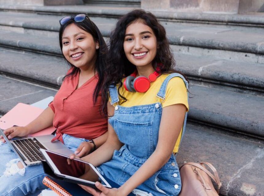Dos mujeres jóvenes sentadas sonrientes y felices en unas escaleras, con sus dispositivos electrónicos en sus manos.