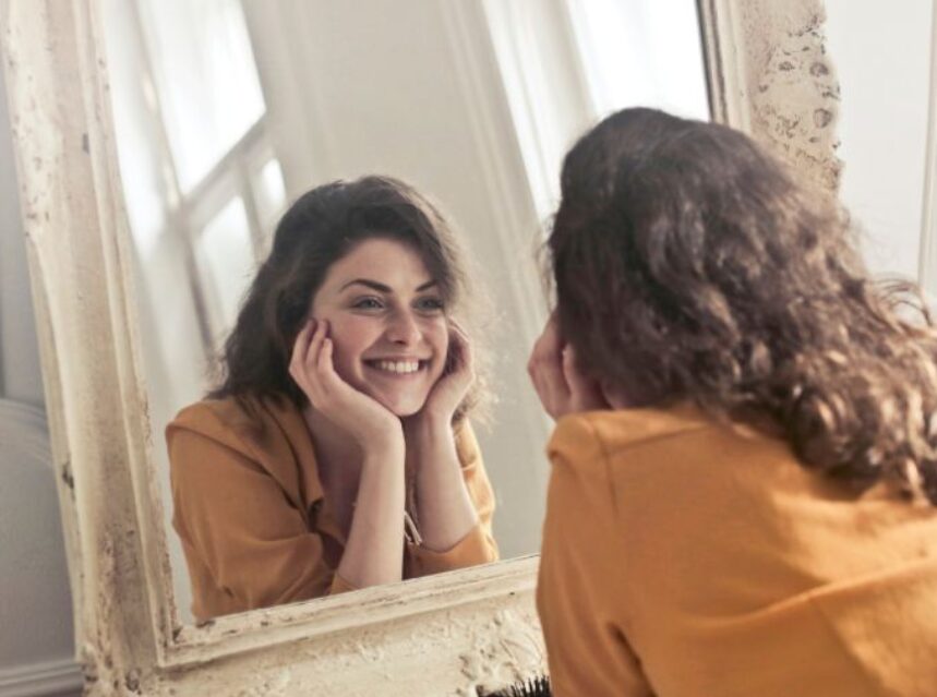 Mujer sonríe frente al espejo en señal de ser amable consigo misma.