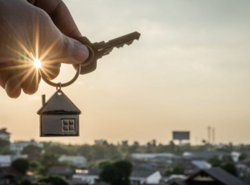 Una mano sostiene unas llaves con llavero de casa, mientras que se filtran los rayos del sol entre los dedos de las manos, a lo lejos se percibe una ciudad.