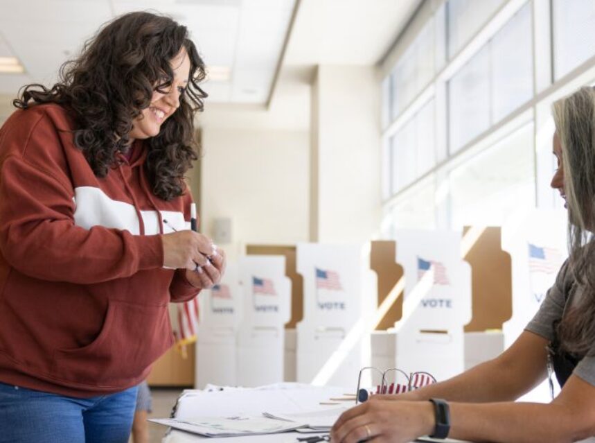 Mujer emite su voto de pie mientras otra mujer en mesa lo recibe.