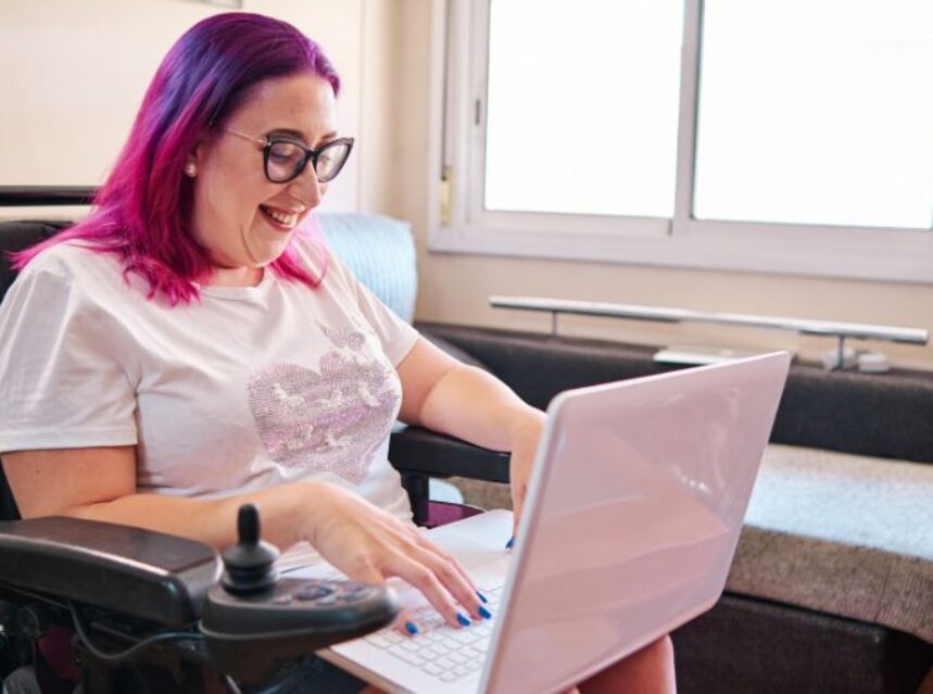 Una joven mujer en su silla adaptada sonríe frente a su laptop.