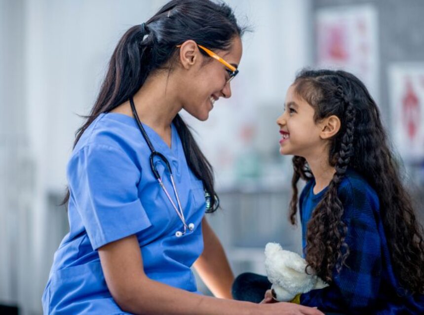 Mujer médico sonríe a paciente niña también sonriente, sobre la camilla de una sala de hospital.