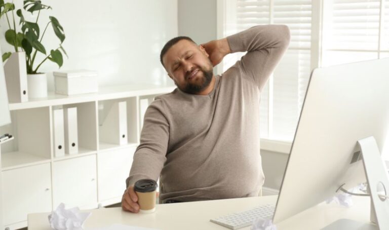 Joven hombre estirándose en señal de flojera, con un café en la mano, frente a su computadora en el escritorio.