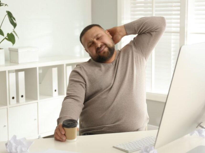 Joven hombre estirándose en señal de flojera, con un café en la mano, frente a su computadora en el escritorio.