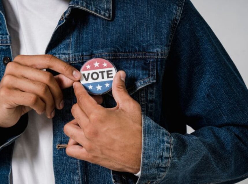 Se ve la parte frontal de una persona con chamarra de mezclilla que se coloca un pin que dice "Vote".