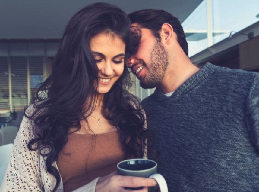 Joven pareja sonriente, ella tiene una taza en su mano y él le susurra algo al oído.