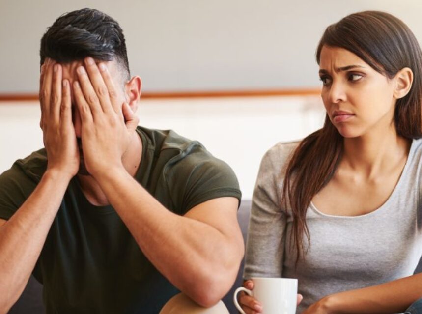 Una pareja está sentada en un sofá, la mujer mira al hombre con una mirada escéptica mientras él cubre su rostro con las manos.