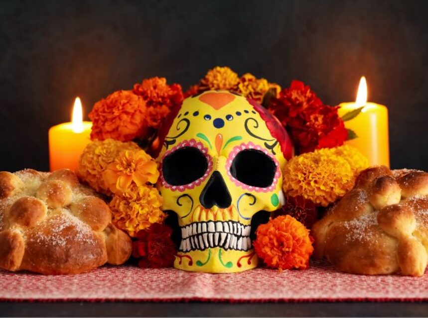 Altar de muertos, dos veladoras, flores, dos panes de muertos y una calavera al centro.