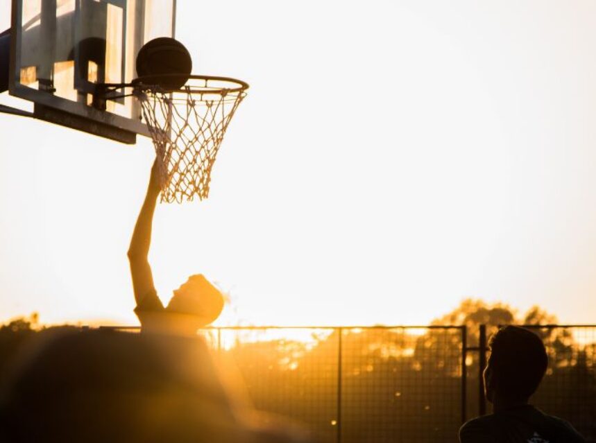 Dos jóvenes juegan básquetbol con un sol resplandeciente de fondo.