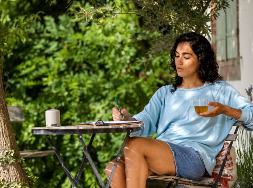 Joven mujer en un jardín, sentada escribiendo un diario y sosteniendo un té en su mano izquierda.