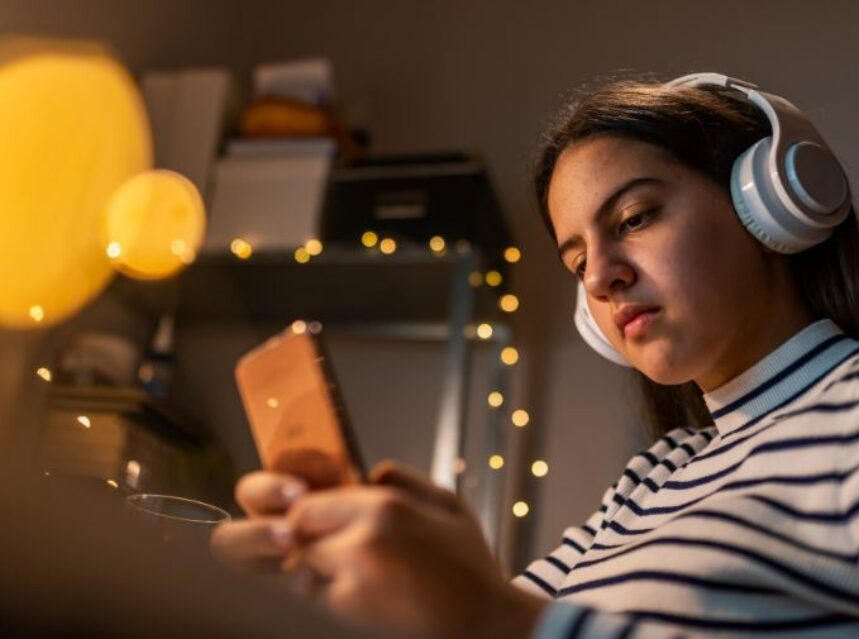 Una joven adolescente mira la pantalla de su celular. Tiene auriculares puestos.