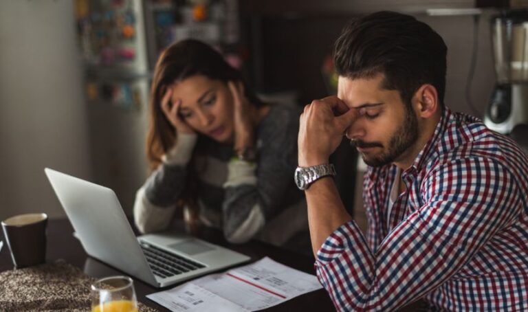 Una pareja está en una mesa con una laptop y unos recibos, con rostros cabizbajos revisan sus cuentas.