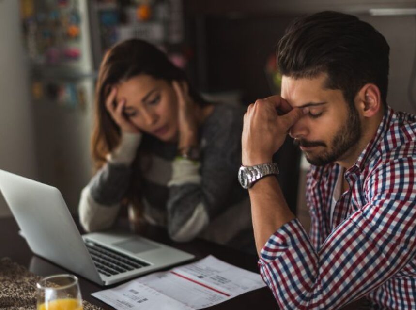 Una pareja está en una mesa con una laptop y unos recibos, con rostros cabizbajos revisan sus cuentas.