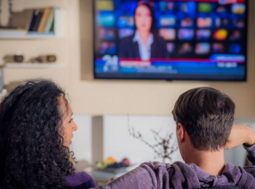 Una pareja sentada en un sofá mira el televisor donde se proyectan las noticias.