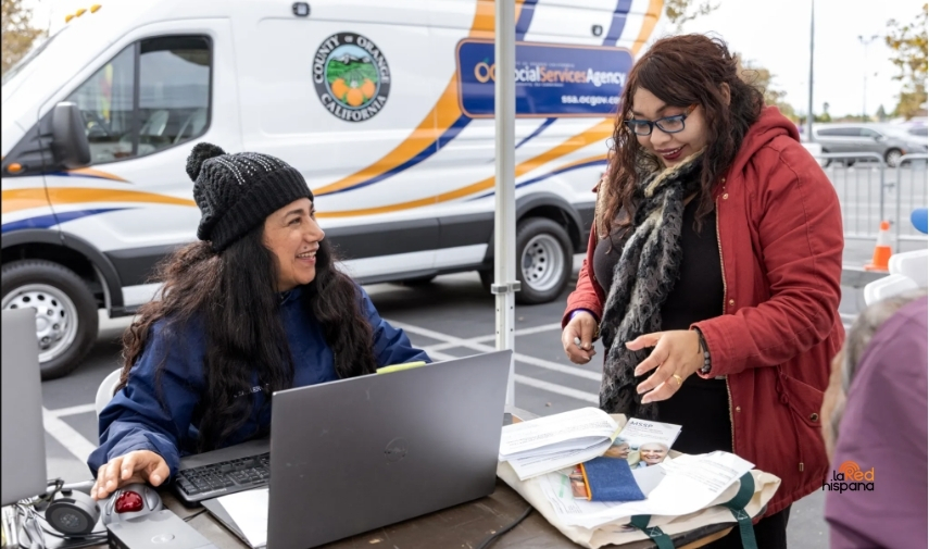Nina Sicaeros, de la Agencia de Servicios Sociales del condado de Orange (izq.), ayuda a Carmen Basu a solicitar el programa CalFresh para su suegra durante un evento de distribución de alimentos con motivo del Día de Acción de Gracias el 22 de noviembre, organizado por la aseguradora de Medi-Cal CalOptima en Orange, California. (Alisha Jucevic for KFF Health News)